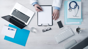 Pictured are the hands of someone in a lab coat writing on a clipboard with computer, glasses, a phone and computers on the desk. The doctor is perhaps calculating his Section 179 deduction for medical equipment.