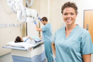 A woman with short, curly hair and wearing scrubs stands in the foreground. In the background a man wearing identical scrubs is taking X-rays of a patient lying on the X-ray table — history of radiology.